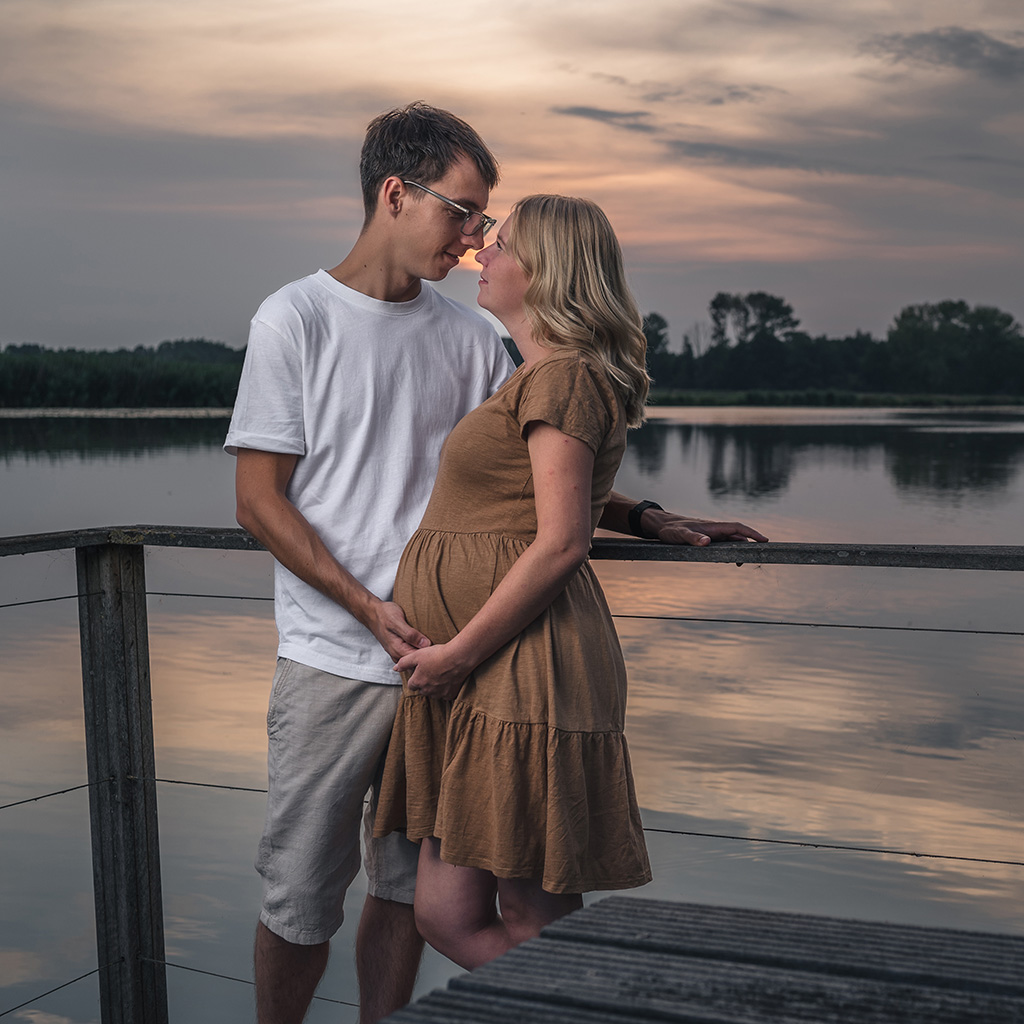 Ein strahlendes Paar während des Sonnenuntergangs beim Babybauchshooting auf dem Steg mit dem Rhein im Hintergrund. Hamminkeln, NRW, Niederrhein.